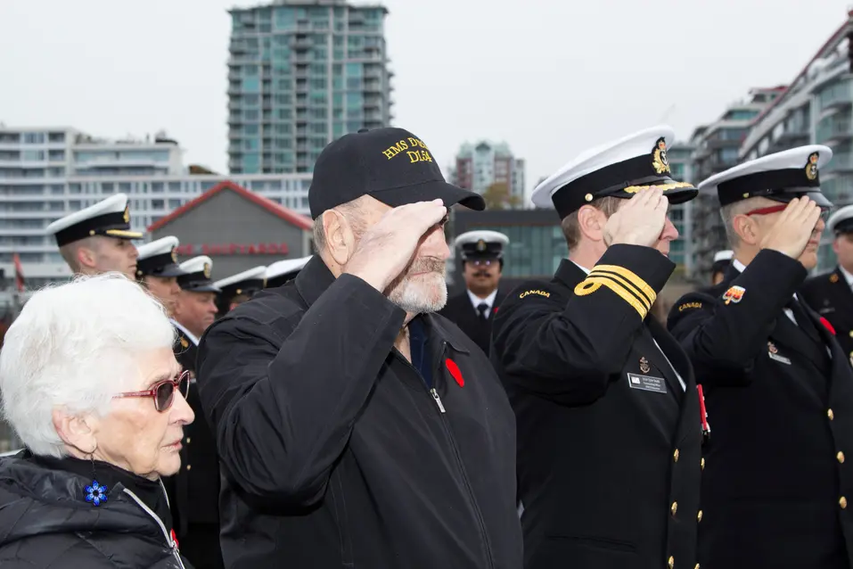 Senior man wearing poppy on his chest is saluting with navy members / Un homme âgé portant un coquelicot sur la poitrine salue avec des membres de la marine.