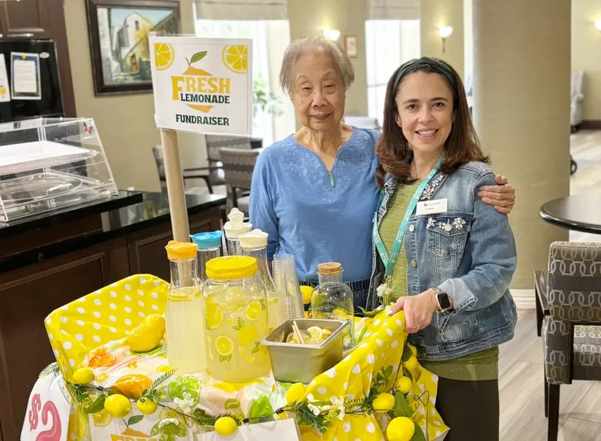Senior woman and young volunteer are posing in front of lemonade stand for fundraiser / Une femme âgée et un jeune bénévole posent devant un stand de limonade pour une collecte de fonds