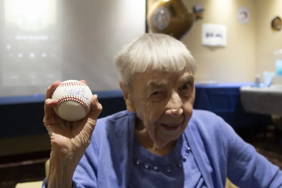 Senior woman smiling big while holding a signed from Blue Jay's team / Femme âgée souriant à pleines dents en tenant un panneau de l'équipe Blue Jay.